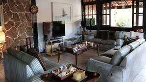 A living room with a stone wall, a television, a sofa, a coffee table, and a window with a view of greenery. - BOAT HOUSE - Charm and Comfort on Geribá Beach (Búzios)