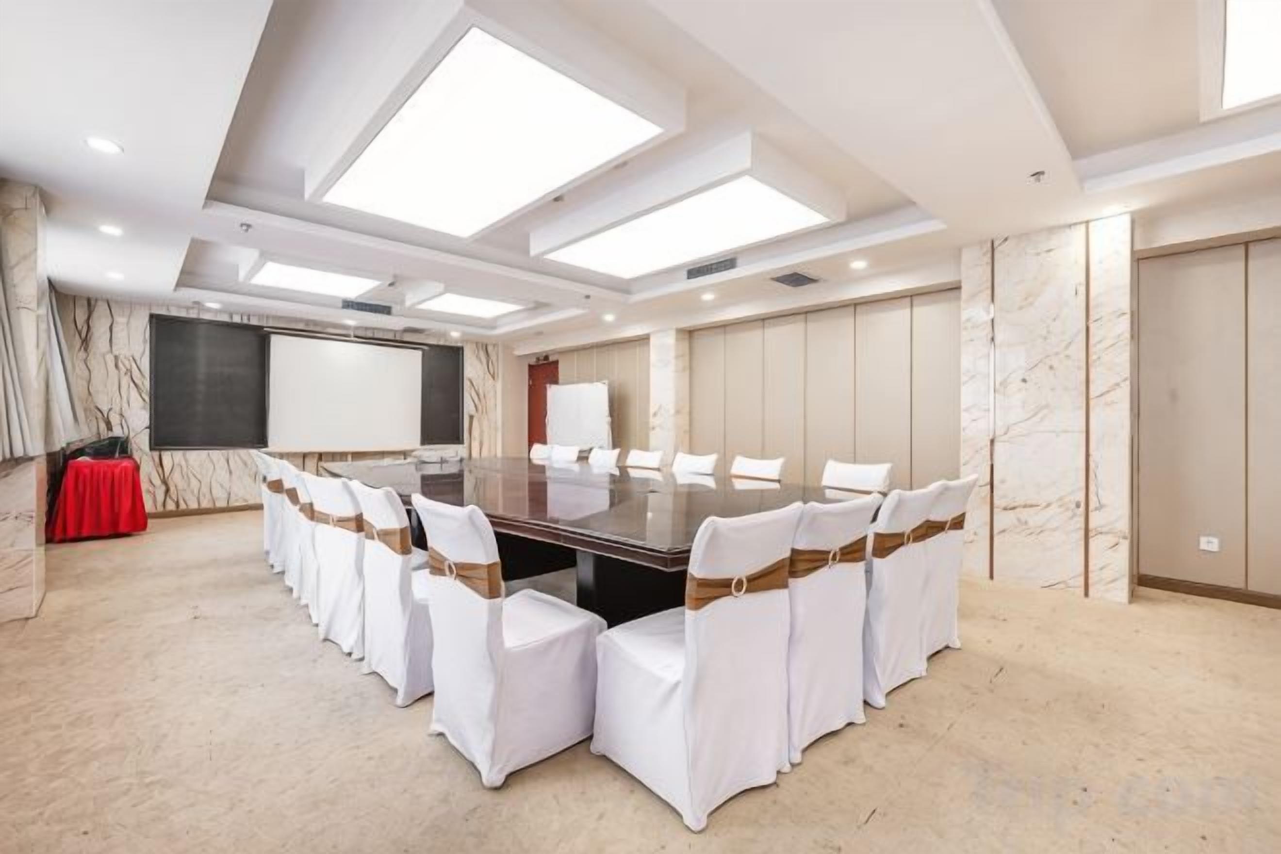 Un salón de conferencias con una gran mesa, sillas blancas y una pantalla de proyector.