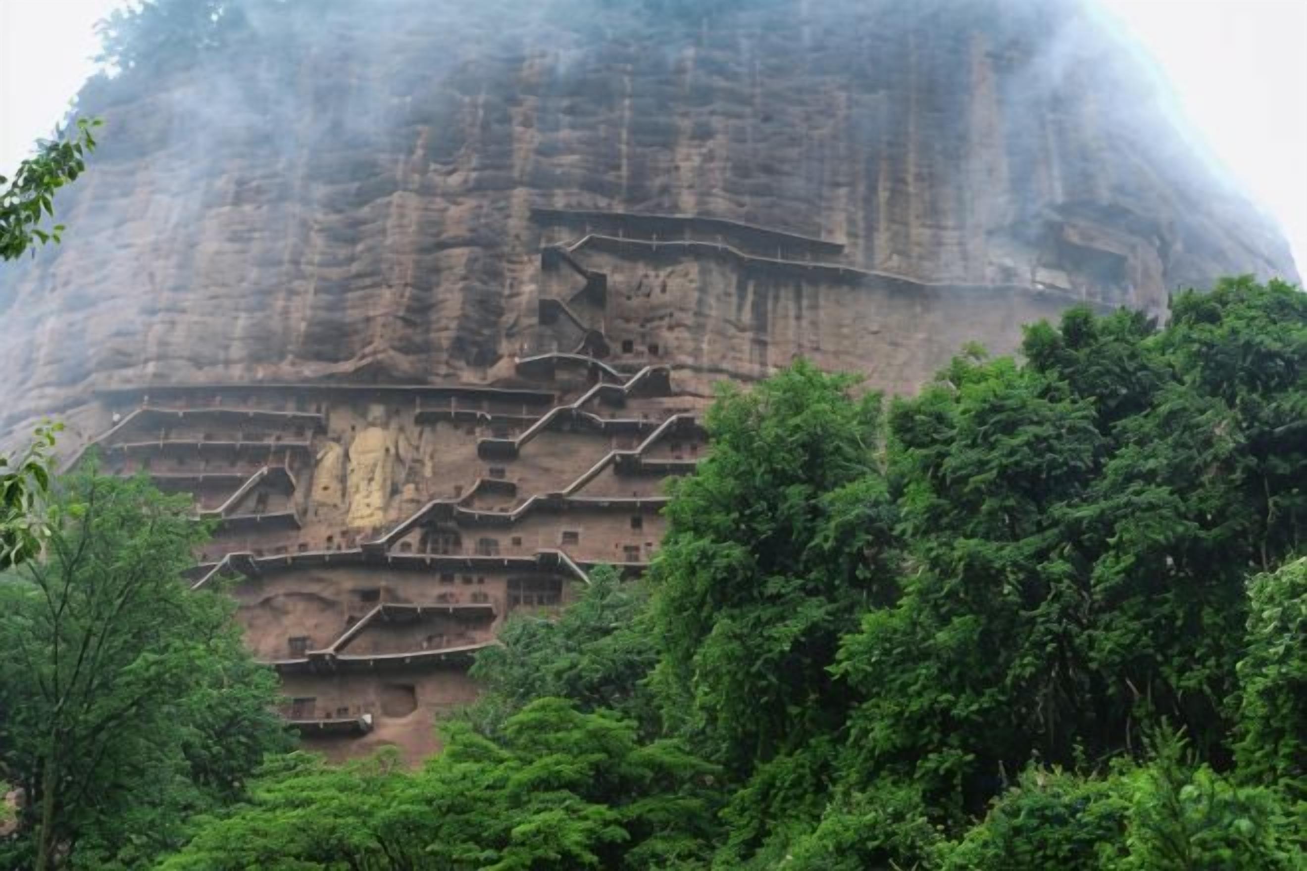 A large rock formation with multiple levels of ancient structures, surrounded by dense green foliage.