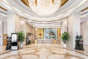 A hotel lobby with a reception desk, a large chandelier, and potted plants.