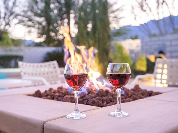 Dos copas de vino sobre una mesa, con un pozo de fuego al fondo.
