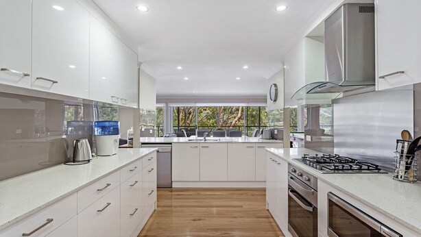 Moderni keittiö, jossa on valkoiset kaapit, suuri keittiösaareke, ruostumattomasta teräksestä valmistetut kodinkoneet ja suuret ikkunat, joista avautuu näkymä puita kohti.