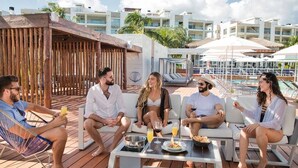A group of people sitting on outdoor lounge chairs at a poolside area with a wooden deck, a pool, and a view of modern buildings in the background. - LA AMADA CANCUN LUXURY CONDO 3 BR STEPS FROM BEACH MARINA (Isla Mujeres)