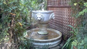Een tuin met een stenen fontein met beeldhouwwerken van leeuwenkoppen, omringd door weelderige beplanting en een houten hek.