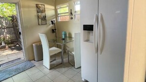 Een kleine keuken met een witte koelkast, een eettafel met stoelen, en een raam dat voor natuurlijk licht zorgt.