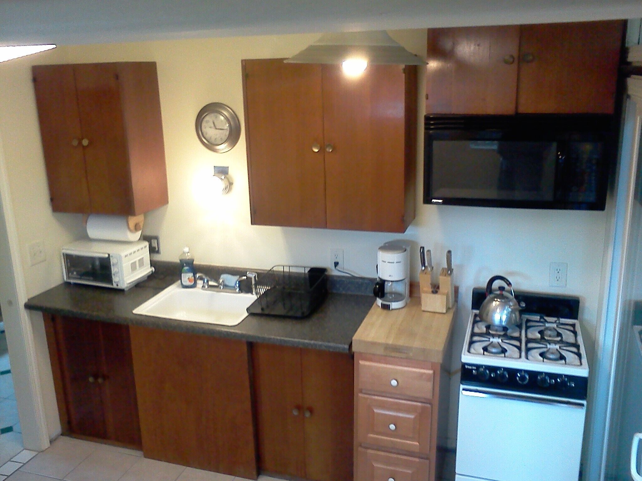 A compact kitchen with wooden cabinets, a microwave, a sink, a stove, and a clock on the wall.