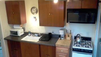 A compact kitchen with wooden cabinets, a microwave, a sink, a stove, and a clock on the wall.