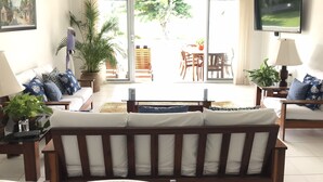 Sala de estar con sofá, televisor y ventilador de techo. Hay una puerta corrediza de vidrio que da a un patio al aire libre con mesa y sillas.