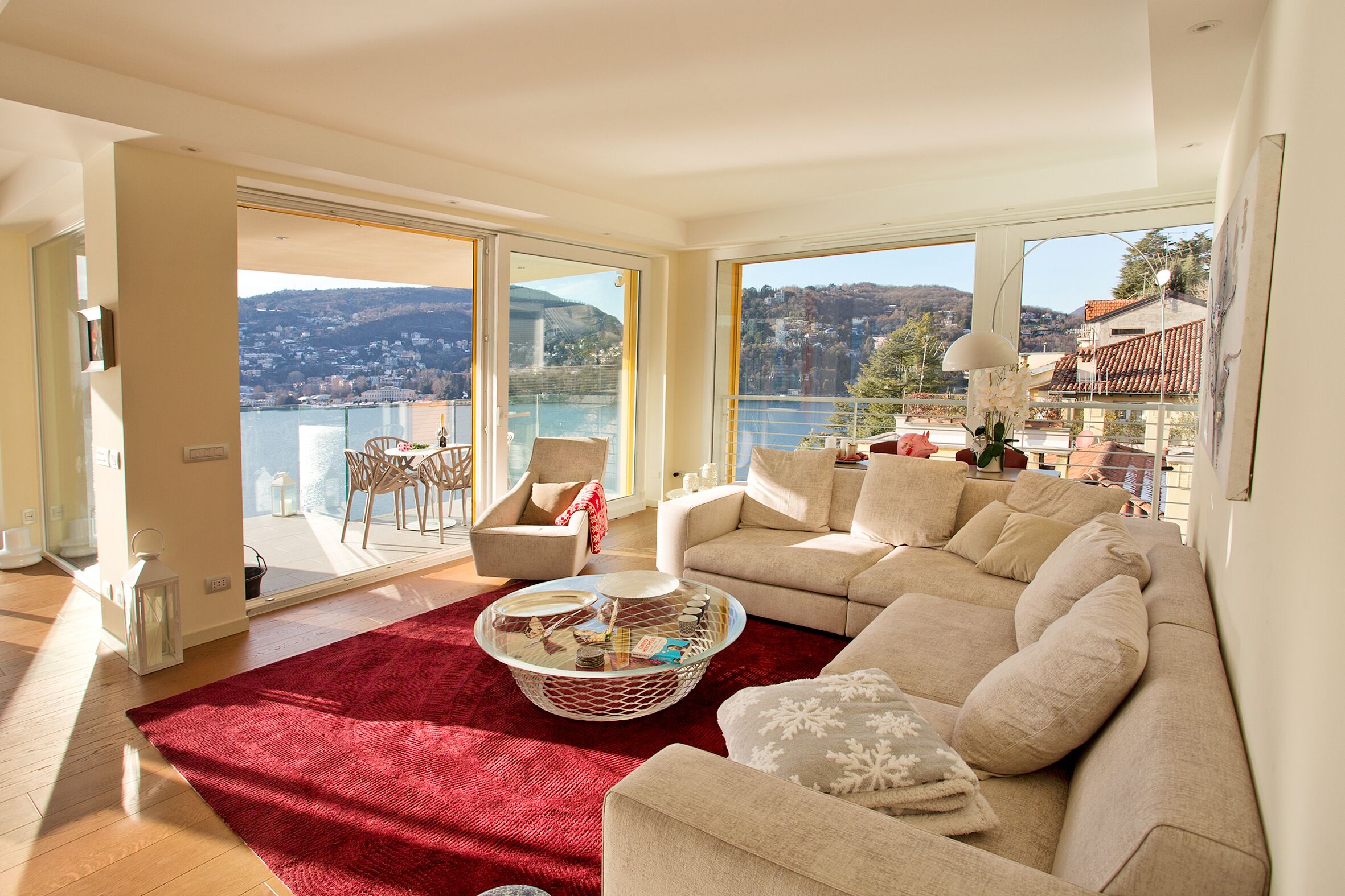 A spacious living room with a large window offering a view of a lake and hills, a red rug, a beige sofa, and a glass coffee table.