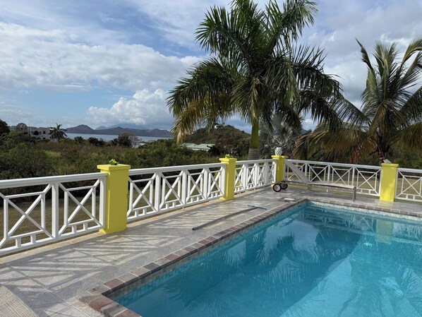 A swimming pool with a white and yellow fence, surrounded by palm trees and a view of mountains and water.