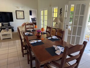 A dining area with a wooden table set for four, surrounded by chairs. There is a television on a stand to the left, a small table with a vase and books, and a lamp on the table.