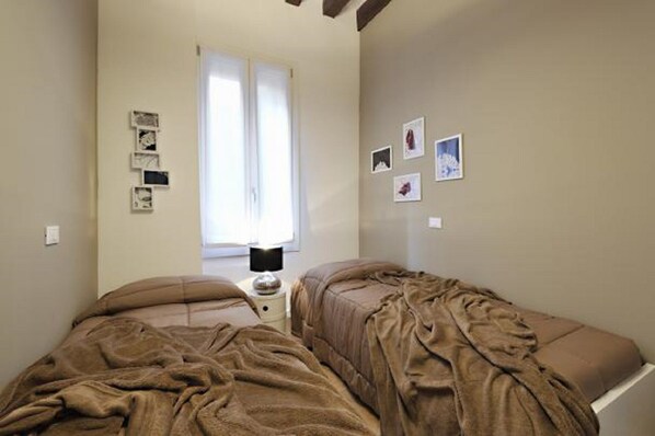 A bedroom with two beds, a window with blinds, and a bedside table with a lamp.