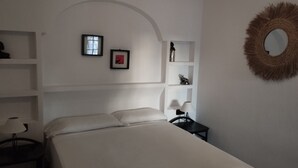 A bedroom with a bed, a nightstand, a lamp, a framed picture, a shelf with a figurine, and a mirror.