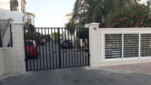 A gated entrance with a security gate, a parking area, and a building with a white facade and a window displaying multiple items.