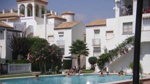 A pool area with multiple swimming pools, surrounded by white buildings with tiled roofs and balconies.