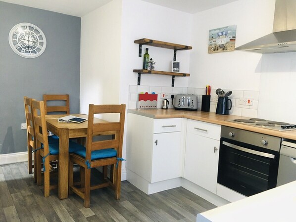 A compact kitchen with a wooden dining table, chairs, a built-in oven, and a small shelf with kitchen items.