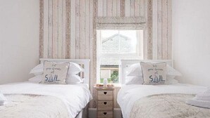Ein Schlafzimmer mit zwei Betten, jedes mit einem Kopfkissen, auf dem 'let your dreams sail' steht. Ein Fenster bietet Blick auf ein Haus, und dazwischen steht ein Nachttisch aus Holz.