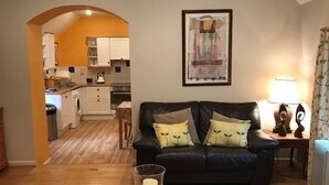 Un salon avec un canapé noir, une table basse en bois et un tapis coloré. On aperçoit une cuisine à travers une arche en arrière-plan.