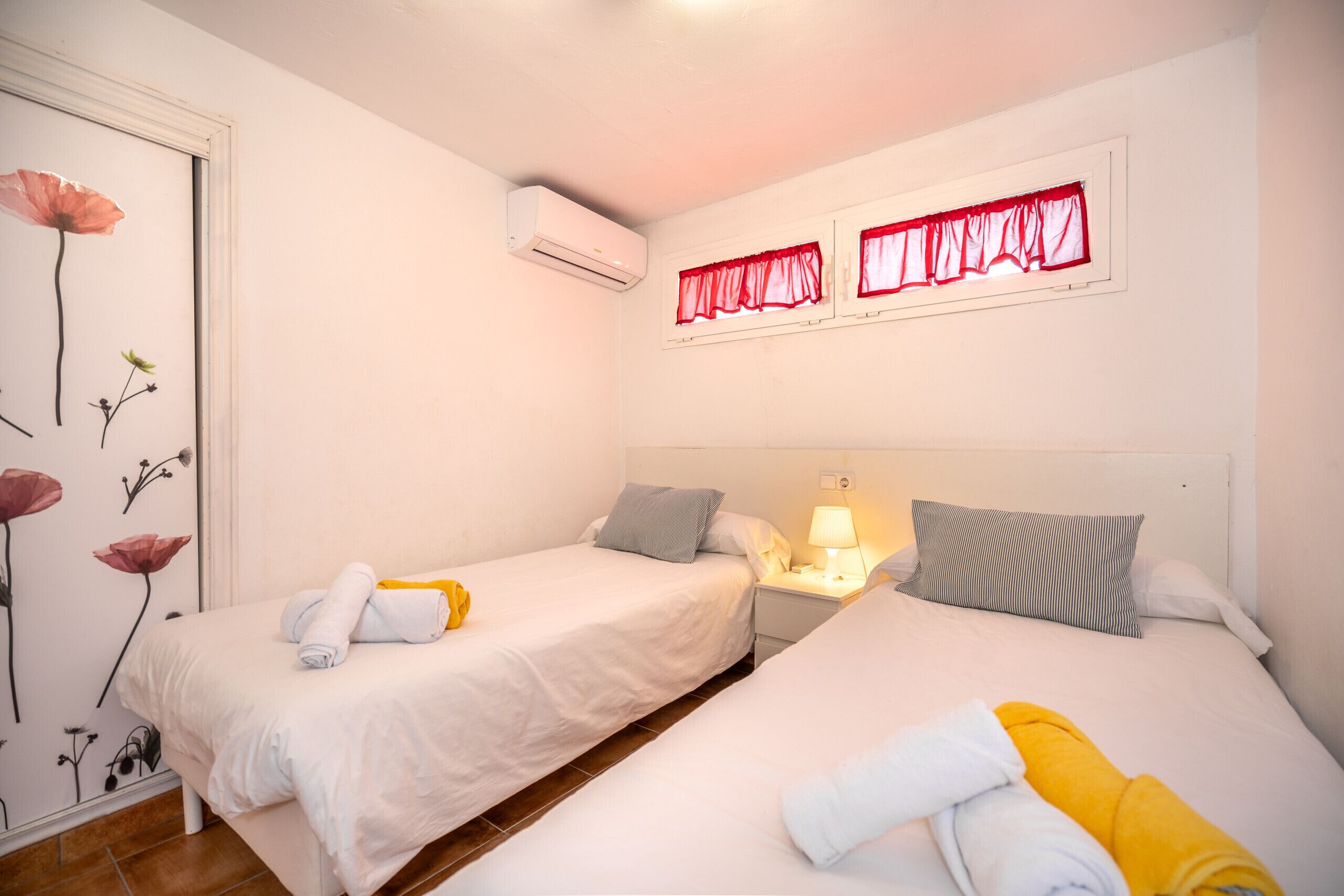 A hotel room with two beds, a nightstand with a lamp, an air conditioner, and a wall with floral wallpaper.