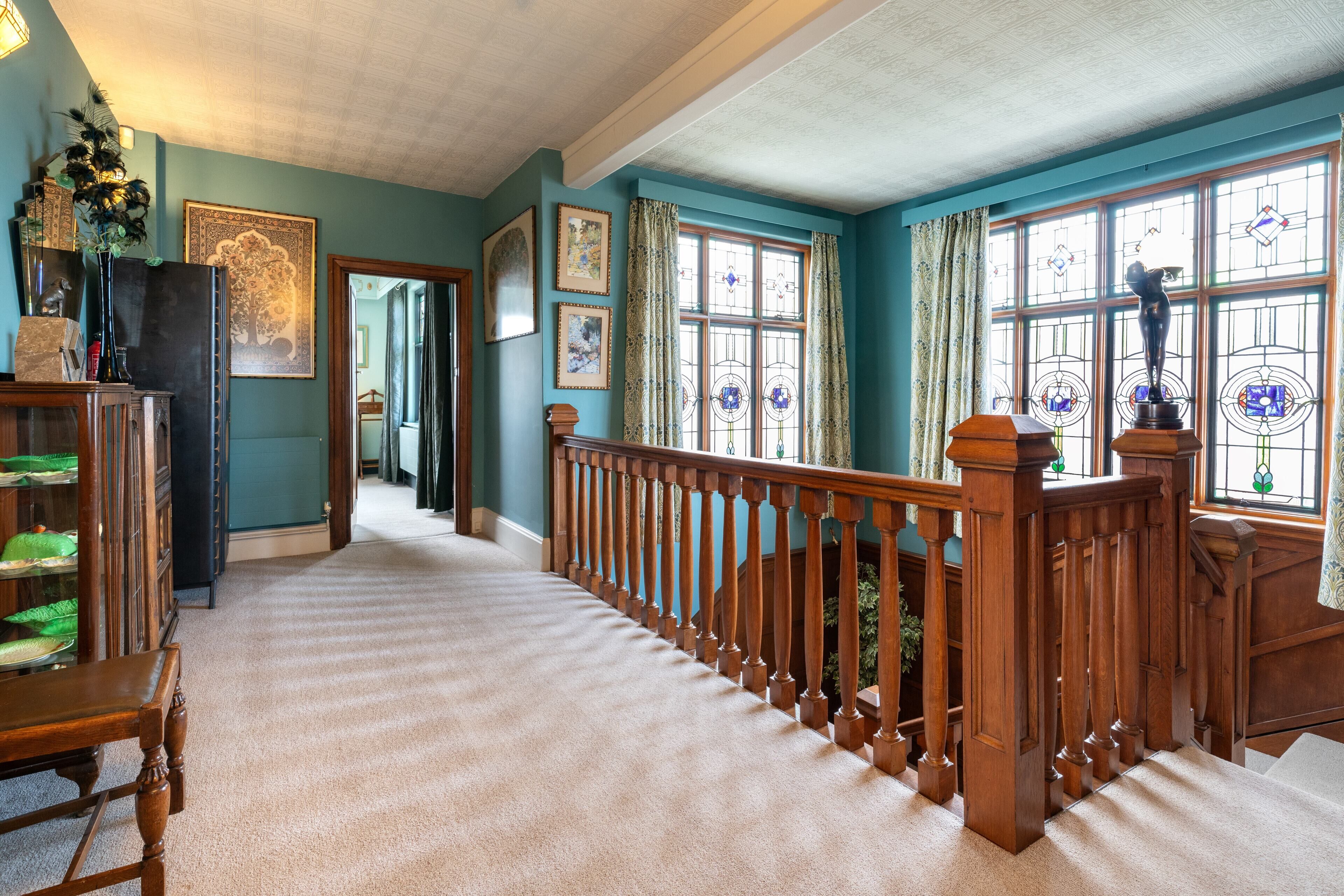 A hallway with a wooden railing, carpeted floor, and a view of a room with a window and curtains.