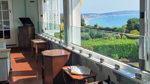 Ein Zimmer mit Meerblick, ein Holzschrank, ein Tisch mit Zeitungen und ein Fenster mit Blick auf das Meer und Grünflächen.