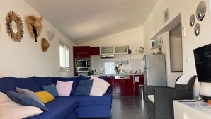 A modern living room with a blue sectional sofa, a dining area with a wicker chair, and a kitchen with red cabinets and stainless steel appliances. - Très bel appartement Porticcio, vue mer sur le golfe d'Ajaccio (Grosseto-Prugna)