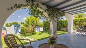 Un espace extérieur aménagé en patio, avec un coin salon couvert, une table agrémentée d’une plante en pot et une vue sur le jardin environnant et les portails bleus.