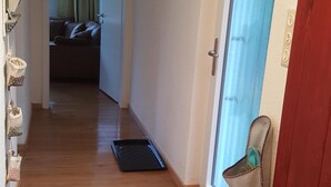 Um corredor com piso de madeira, porta com moldura vermelha, janela com cortinas azuis e um tapete pequeno com padrão floral.