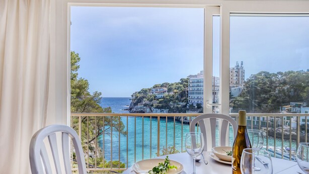A table set for two with a bottle of wine and glasses, overlooking a coastal view with buildings and cliffs. - Drac 5 by dracmallorca (Cala Santanyi)