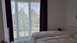 Quarto com cama, janela com cortinas, piso de madeira e vista para árvores e edifícios.