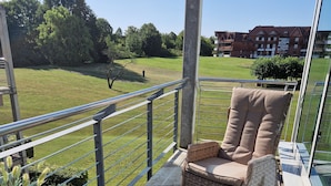 Varanda com móveis de vime, uma mesa pequena e vista para um campo verde e edifícios ao longe.