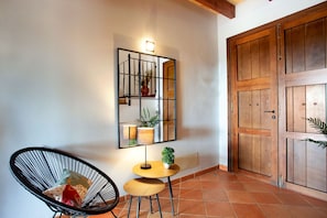 A room with a wooden door, a chair, a small table with a lamp, and a wall-mounted mirror with a plant. - Bassa Seca by dracmallorca (Santanyi)