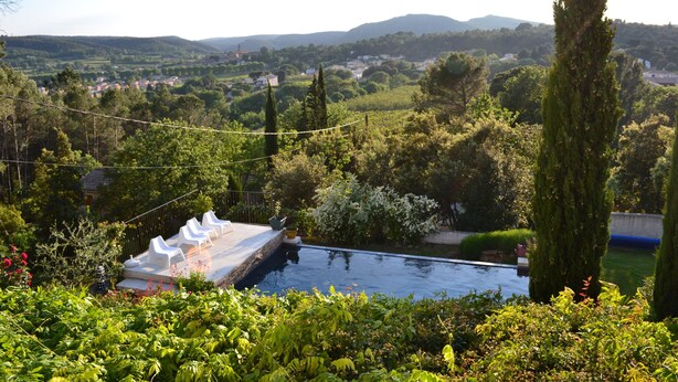 A pool with a deck, surrounded by lush greenery and tall trees. - Beautiful house with private heated infinity pool 16 km from Uzes (Connaux)