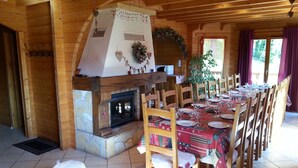 Interior de una cabaña de madera con chimenea, mesa de comedor para seis personas y un ventanal con cortinas.