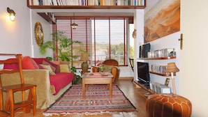 Una sala de estar acogedora con un sofá, una mesa de centro de madera, una estantería y un ventanal con persianas.