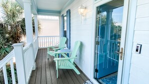 Un porche cubierto con suelo de madera, sillas Adirondack azul claro y una puerta de cristal que da a una piscina.