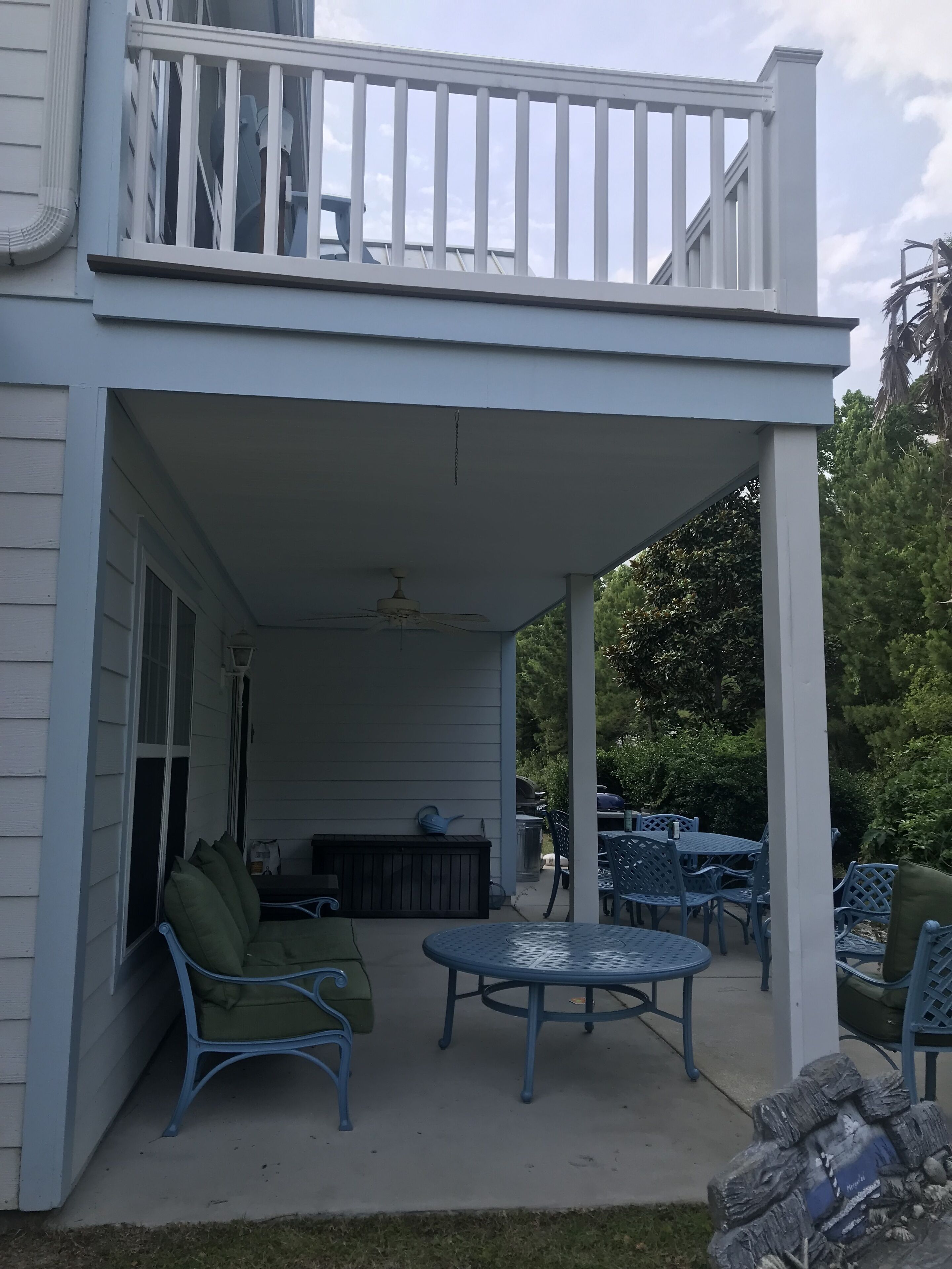 A covered patio area with a balcony, outdoor furniture, and a view of the surrounding greenery.