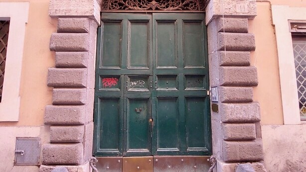 Une porte verte à deux battants, ornée de sculptures complexes, flanquée de piliers en pierre et donnant sur un pavé de pierres.