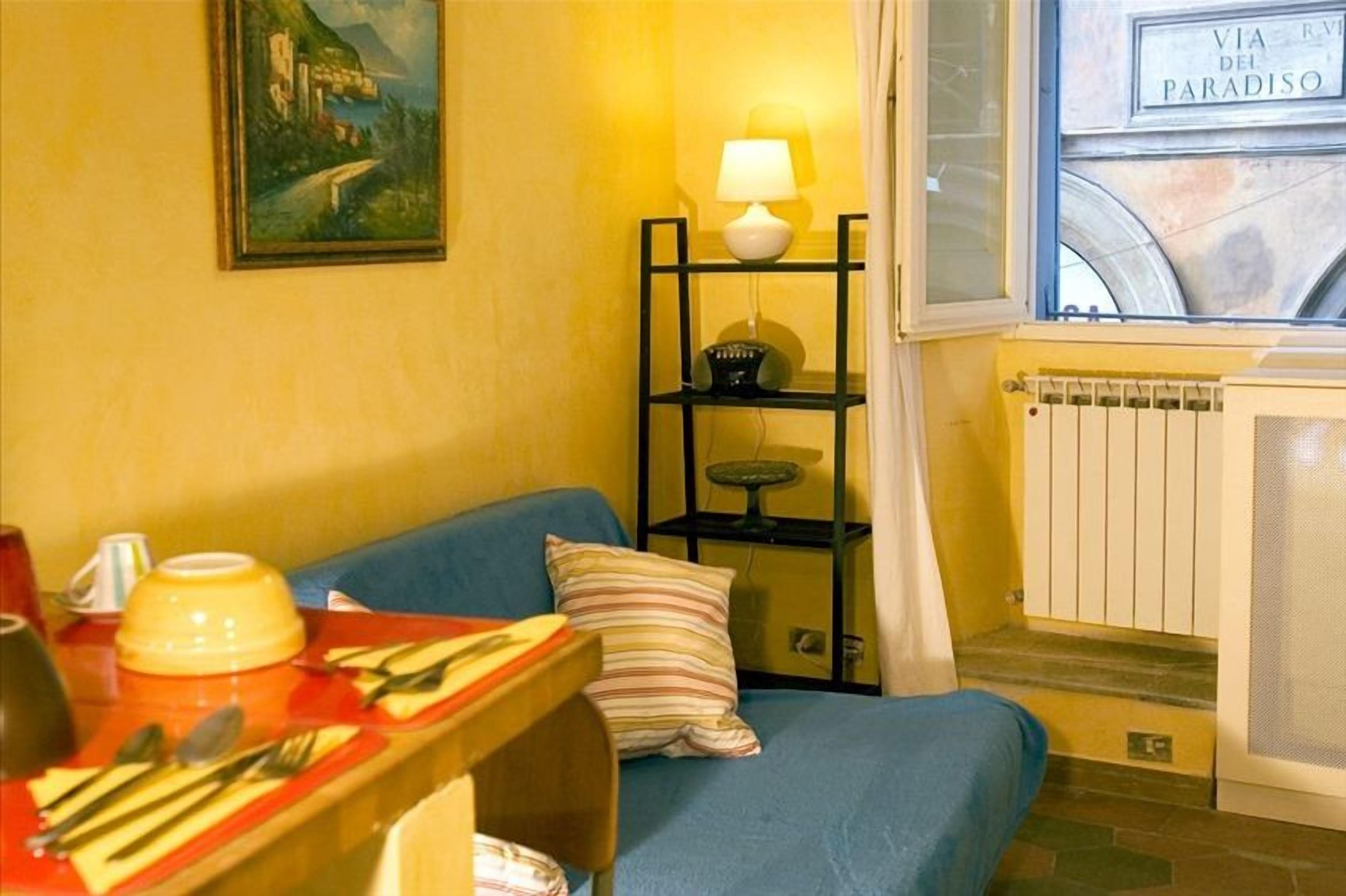 A cozy room with a blue sofa, a small table with a lamp, and a window with a view of a building labeled "VIA DEL PARADISO".
