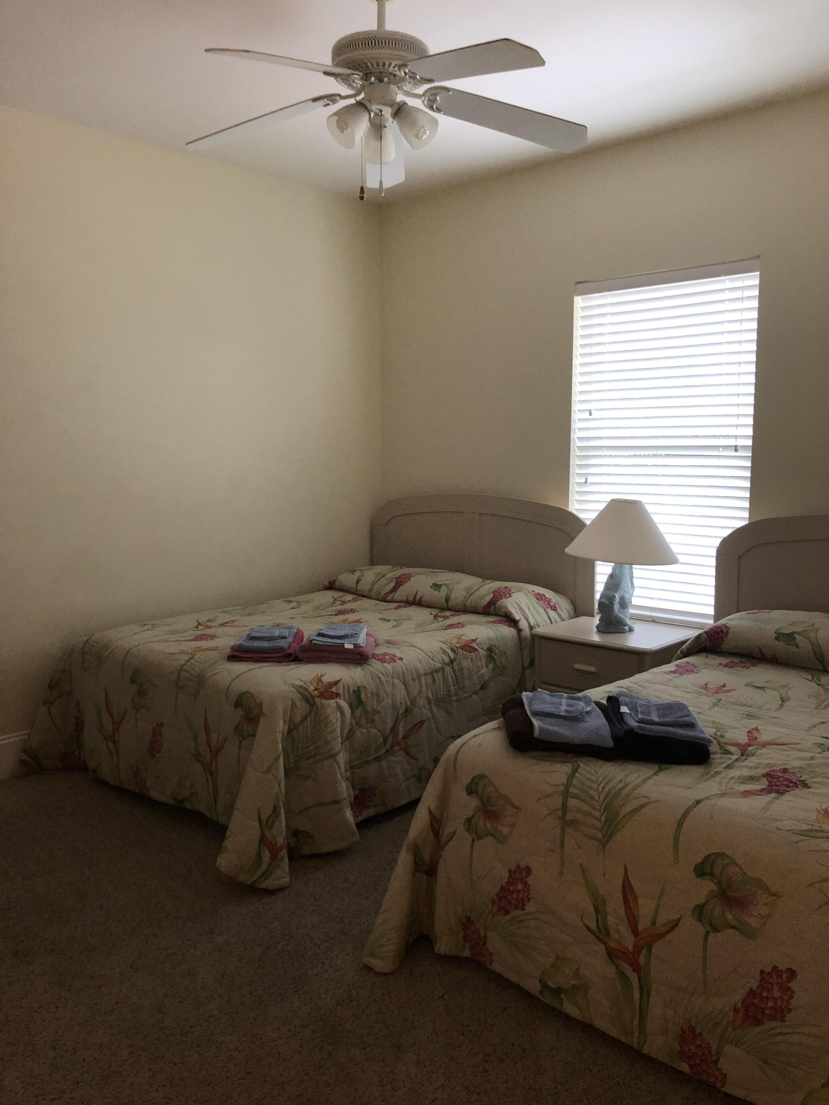A bedroom with two single beds, a ceiling fan, a window with blinds, and a bedside table with a lamp.