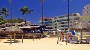 A beach with palm trees, beach chairs, and a building with multiple floors in the background. - Experience the Best of Bucerias Beach Front Living (Bucerias)
