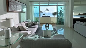 A modern living room with a sofa, a glass coffee table, and a view of the ocean through large windows. - Experience the Best of Bucerias Beach Front Living (Bucerias)