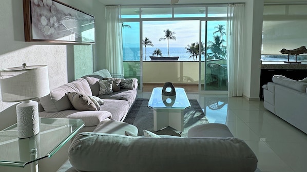 A modern living room with a sofa, a glass coffee table, and a view of the ocean through large windows. - Experience the Best of Bucerias Beach Front Living (Bucerias)