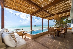 Une piscine à débordement avec une terrasse en bois, un canapé et des chaises de salle à manger, le tout offrant une vue sur l’océan.