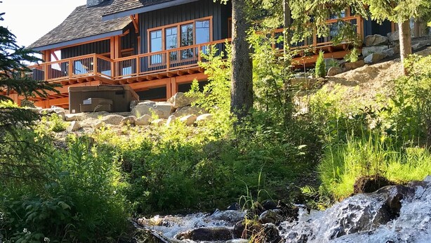 A large wooden house with multiple balconies, surrounded by lush greenery and a flowing stream. - Exceptional Ski-in/Walk-out Home - 4100 Sft (Golden)