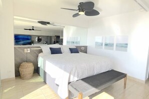 A modern bedroom with a large bed, a bench, a ceiling fan, and a view of the ocean through the window.