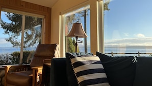 A cozy room with a leather chair, a dark sofa with striped pillows, a lamp, and large windows offering a view of the sea and trees. - Top-Rated Waterfront Retreat Near Port Townsend (Nordland)