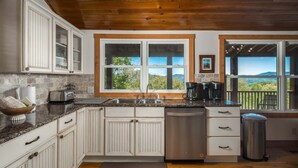 A kitchen with white cabinetry, a granite countertop, a stainless steel dishwasher, and a large window with a view of the outdoors.