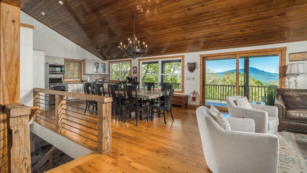 A spacious kitchen with wooden ceiling, a dining area with a round table, and a living room with a sofa and a view of the mountains.