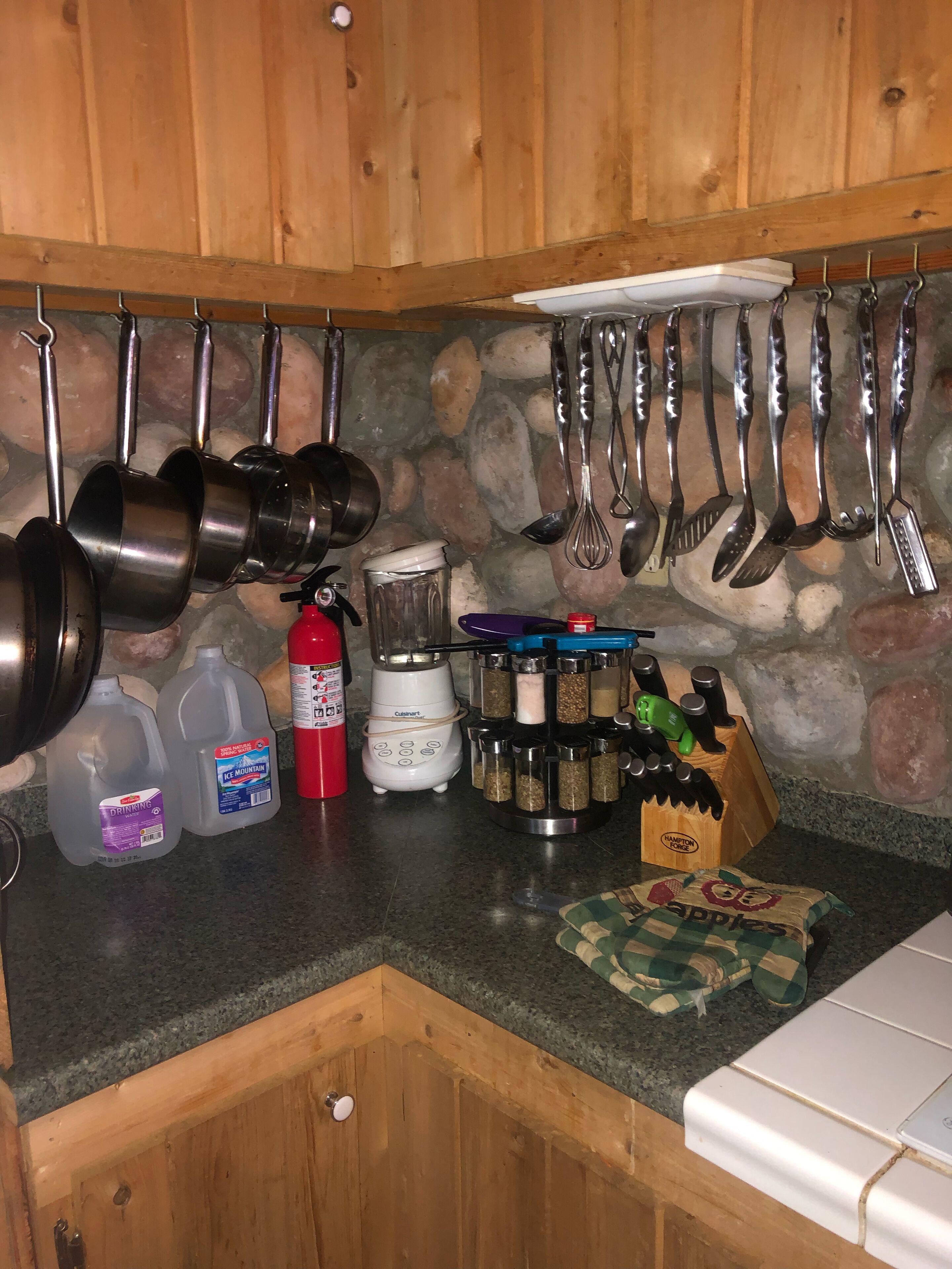 A kitchen with wooden cabinets, a stone backsplash, and various utensils hanging above the counter. There are two bottles of water, a red fire extinguisher, and a blender on the counter.